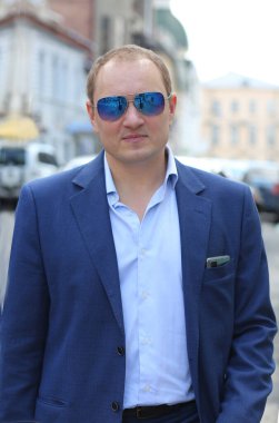 Mavi takım elbiseli, elinde evrak çantası tutan güneş gözlüklü, kendinden emin beyaz bir gencin portresi. Arka plan şehrin hareketli iş merkezidir.. 