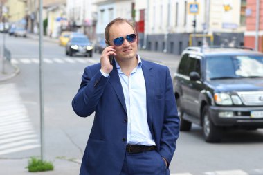 Mavi takım elbiseli ve güneş gözlüklü beyaz bir adamla afişte iş görüşmeleri için akıllı bir telefon var. İş, iş. Arka plan - şehirdeki gürültülü merkez.