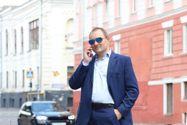 Mavi takım elbiseli ve güneş gözlüklü beyaz bir adamla afişte iş görüşmeleri için akıllı bir telefon var. İş, iş. Uzayı sola kopyala