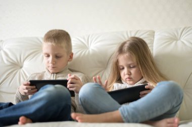 İki küçük çocuk ellerinde tablet tutarak evde çizgi film izliyorlar. Ağabey ve abla kanepede oturmuş dijital cihazla eğleniyorlar..