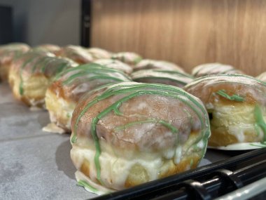 Kremalı ve parlak jöleli taze pişmiş çörekler, yeşil krema çizgileriyle süslenmiş. Fırın ortamında parşömen kâğıdı üzerine dizilmiş. Sığ alan derinliği, sanatsal hamur işi üretim konsepti.