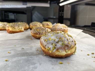 Süslü, beyaz kremalı ve şam fıstıklı bir sıra jöleli çörek. Minimalist kompozisyon, seçici odaklanma ve doğal ışık. Taze tatlı, modern fırın iş akışı ve tatlı yemek fotoğrafçılığı..