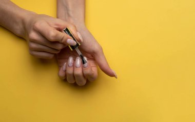 Bir kadının yakın plan fotoğrafı sarı arka planda, üst manzaralı, lakteriyle tırnaklarını boyuyor. Manikür konsepti, düz yatak