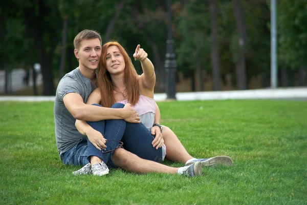 Couple sitting on the grass in the park. Woman shows a finger gu — Stock Photo, Image