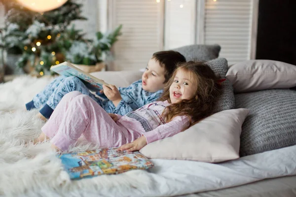 Küçük çocuk ve pijamalı kız yatakta uzanıp Noel ağacının yanında ışık saçan kitaplar okuyorlar. Tatlı Noel çocukları.