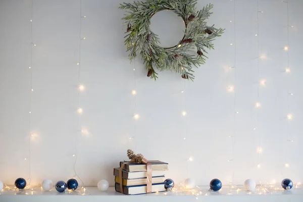 Kurdeleli kitaplar, Noel baloları, Noel çelenkleri ve beyaz duvarın yanındaki Noel ışıkları.