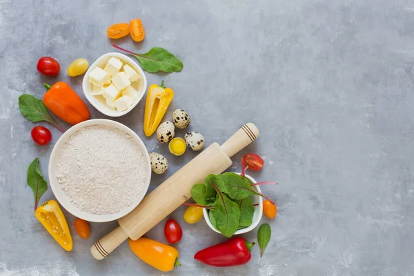Ingredients for cooking vegetable pie with copy space top view. Fresh colorful vegetables, quail eggs, butter, whole grain flour and rolling pin on a grey background. Bakery background