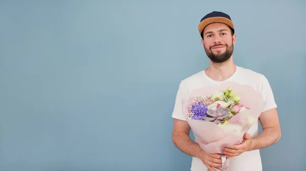 Delivery man in a cap and white t-shirt with beautiful bouquet on a blue background. Flowers delivery concept. Banner with man with flowers