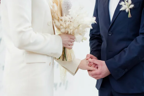 花嫁と新郎の交換結婚指輪 花嫁でベージュの結婚式のスーツで花束ドライ花と新郎でダークブルーのスーツで彼らの結婚式の日 — ストック写真