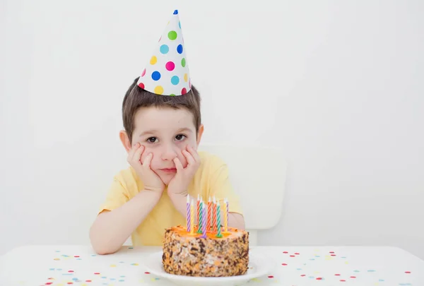 Sad toddler caucasian boy in colorful birthday hat sitting near birthday cake with blowing candles on a white background, waiting for party