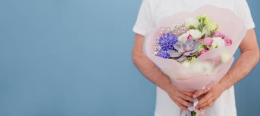 Beyaz tişörtlü, mavi arka planda güzel buketli bir teslimatçı. Çiçek teslimat konsepti. Çiçekli adamla bayrak