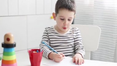 Küçük beyaz çocuk evde resim çiziyor. Aydınlık odada beyaz sandalyede oturan ve beyaz masaya resim çizen çocuk.