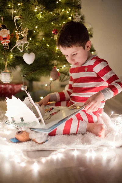 Çizgili pijamalı beyaz çocuk Noel ağacının yanında oturuyor. Dekorasyon ve ışıklarla dolu samimi bir Noel arifesi..
