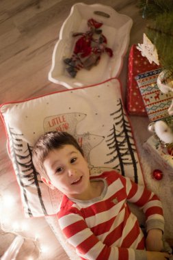 Çizgili pijamalı beyaz çocuk Noel ağacının altında yatıyor. Dekorasyon ve ışıklarla dolu samimi bir Noel arifesi..