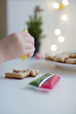 Tatlı çocuk Noel kurabiyelerini Noel ağacının yanındaki beyaz bir masada renkli kremalarla süslüyor. Noel yemeği konsepti