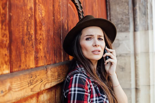 Mujer joven con un sombrero al lado de una vieja puerta de madera hablando en cel — Foto de Stock