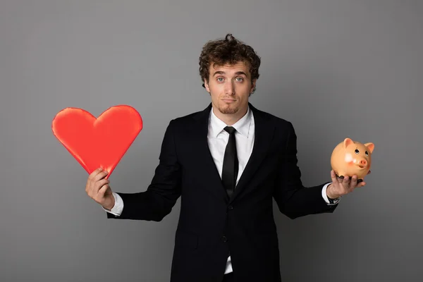 Bonito homem no shuit segurando um vermelho coração e um porquinho banco em ha — Fotografia de Stock