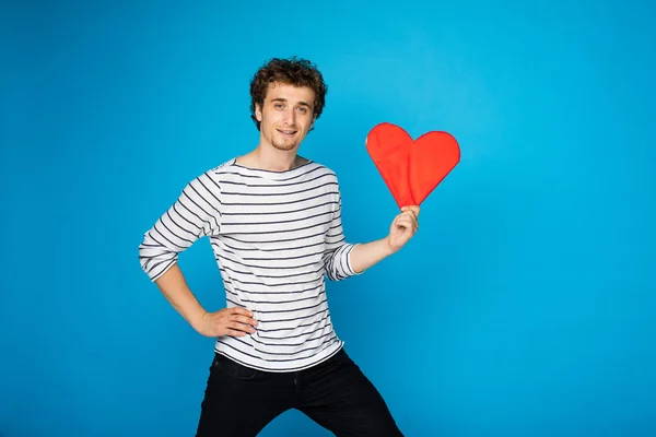 Young curly man with red heart on blue background — Stock Photo, Image