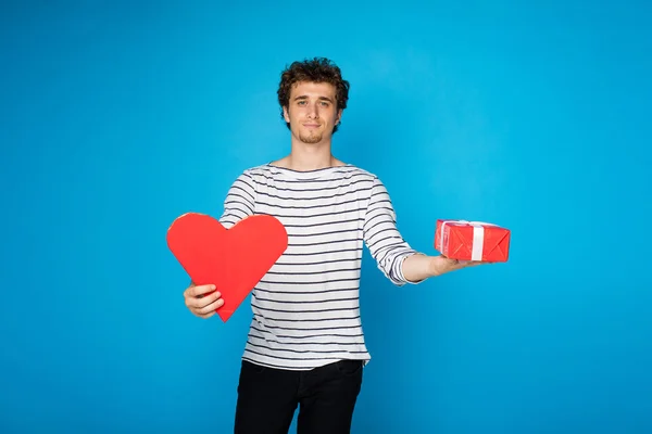Young curly man with red heart and a gift on blue background — Stock Photo, Image