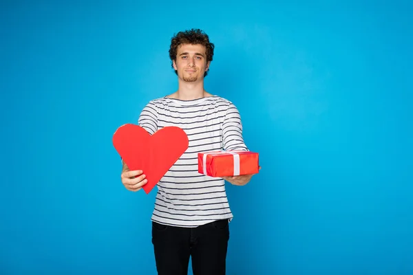 Junger lockiger Mann mit rotem Herz und einem Geschenk auf blauem Hintergrund — Stockfoto