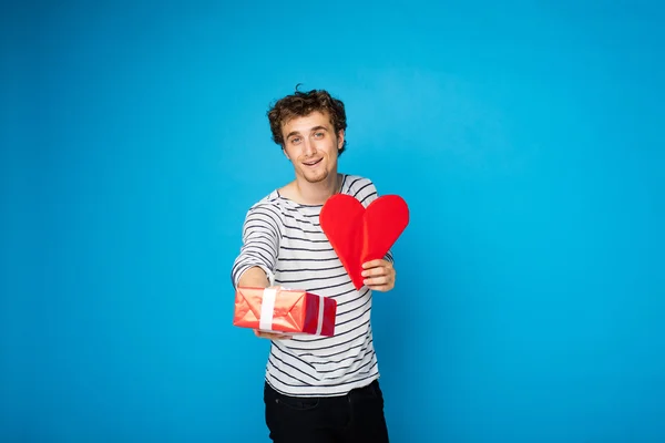 Young curly man with red heart and a gift on blue background — Stock Photo, Image