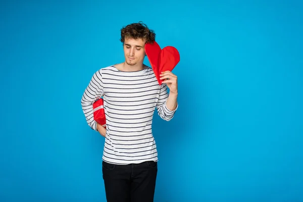 Giovane uomo riccio con cuore rosso e un regalo su sfondo blu — Foto Stock