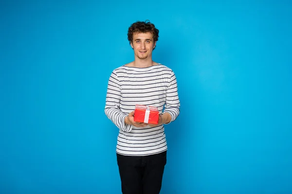 Joven hombre rizado con corazón rojo y un regalo sobre fondo azul — Foto de Stock