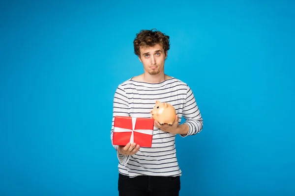 Handsome curly young man with a red gift and a piggy bank on blu — Stock Photo, Image