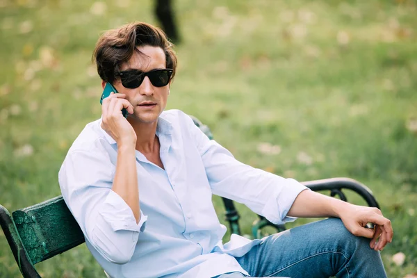 Handsome adult man sitting on bench and talking at cellphone — Stock Photo, Image