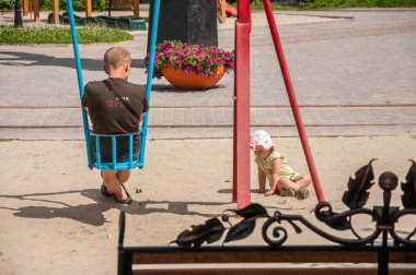 Yetişkin bir adam salıncakta sallanırken çocuğunun kumlarda oynamasını izliyor.