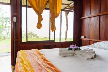 Beyaz havluyla sarılı bir otelin ahşap odası. Yatağın üstüne çiçekler ve perdeli büyük bir pencere.