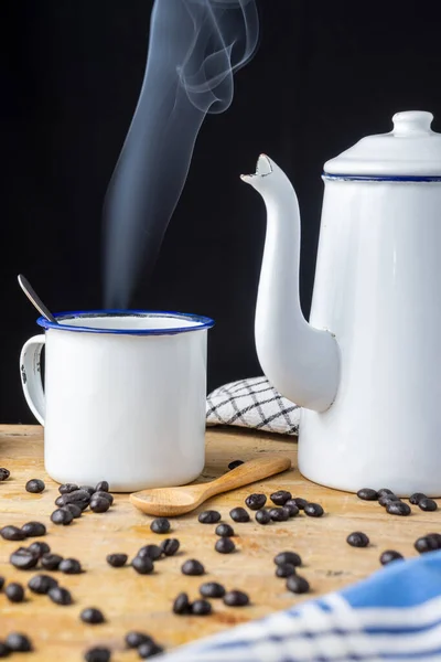 Eski kahve demliği ve beyaz bardakla yakın plan, dumanlı, kahve çekirdeği ve kaşık ahşap masa ve siyah arka plan, dikey.