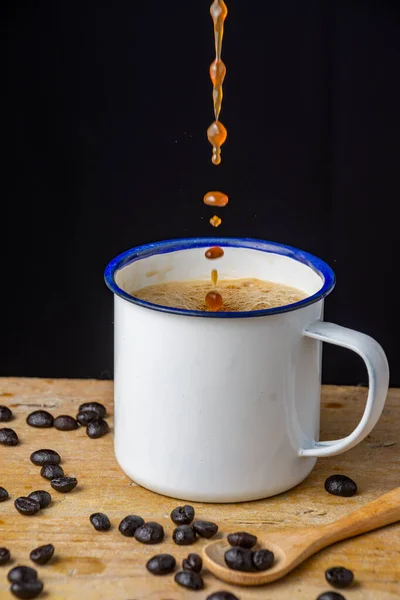 Close-up white cup with falling coffee, coffee beans and spoon on wooden table and black background, vertical