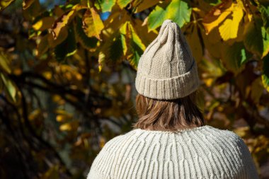 Sonbahar ağaçlarının arka planında süveter ve beyaz şapkalı, yatay bir kadın.