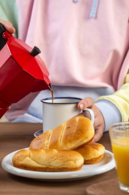 Kazak giyen bir kadının ellerinin üst görüntüsü, kırmızı kahve demliğinden kahve servisi, kahvaltıda rulo ekmek, dikey olarak.