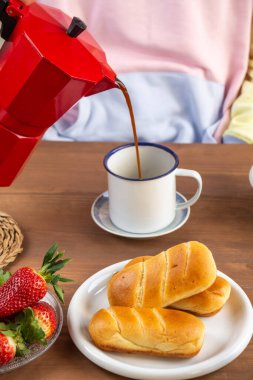 Spor kıyafetleri giyen, kırmızı kahve makinesinden beyaz fincana kahve servisi yapan, kahvaltı masasında çörekler ve meyveli, seçici odaklı, dikey...