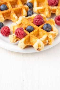 Tabaktaki ahududu ve yaban mersinli waffle 'ların üst görüntüsü, seçici odak, beyaz masa, dikey, kopyalama alanı