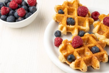 Ahududu ve yaban mersinli waffle, ahududu ve yaban mersinli kase, beyaz masaya seçici bir odak noktası,