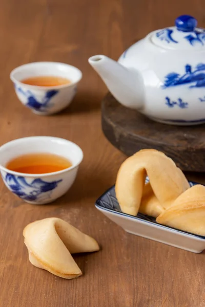 Top view of fortune cookies on a plate, teapot and Chinese cups with tea, on wooden table, with selective focus, vertical