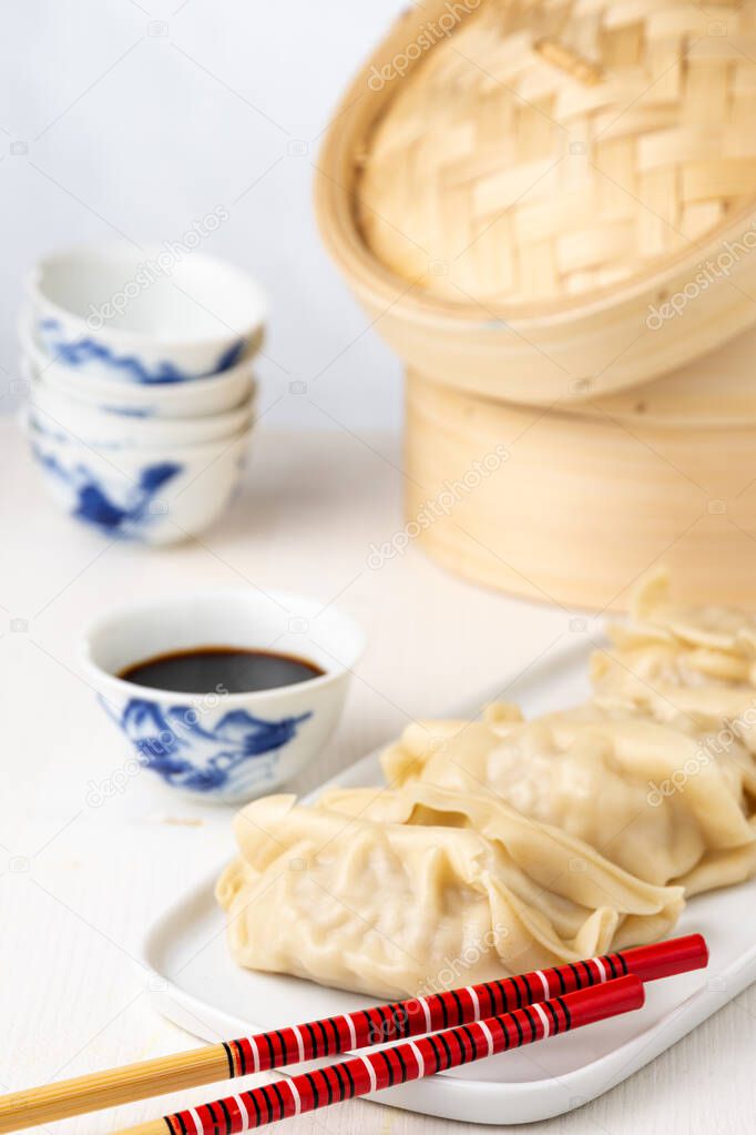 Top view of white plate with gyozas and chopsticks with soy bowl and steamer on white background, vertical