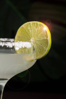 Yarım bardak margarita kokteyli, limon dilimi ve tuzlu, siyah arka plan yeşil yapraklı, dikey
