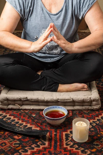 Olgun bir kadının vücudu rahat kıyafetlerle meditasyon yaparken, gri bir yastık üzerinde tütsü, bir fincan çay ve mum, dikey olarak.