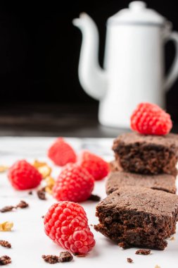 Ekmek kırıntıları, cevizler, kırmızı ahududular ve beyaz kahve demliği olan çikolatalı brownie 'ye yakın plan, beyaz mermer masa, siyah arka plan, dikey...