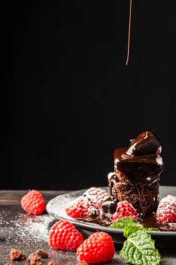 Browni, ahududu ve nane yapraklı tabağın yakın plan resmi. Düşen çikolata şurubu, ahşap masa, seçici odak, siyah arka plan, dikey.