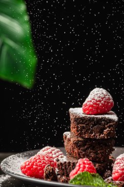 Tabağın kapağı browni, ahududu ve nane yapraklı ve şekerli, seçici odaklı, siyah arkaplan ve yeşil yapraklı, dikey