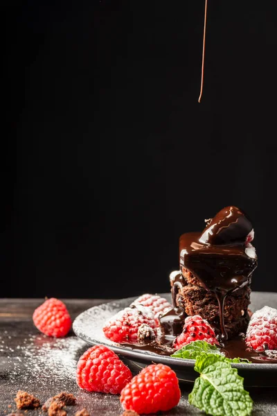 Browni, ahududu ve nane yapraklı tabağın yakın plan resmi. Düşen çikolata şurubu, ahşap masa, seçici odak, siyah arka plan, dikey.