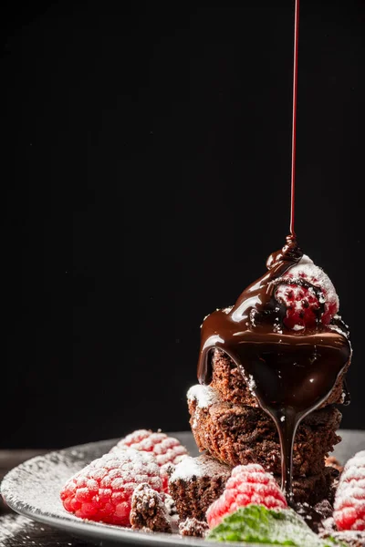 Tabağın kapağı browni, ahududu ve nane yapraklarıyla kapanıyor çikolata şurubu dökülüyor, seçici odak, siyah arka plan, dikey, kopyalama alanı ile birlikte.