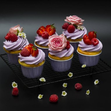 Gül ve papatya çiçekleriyle süslenmiş ahududu ve çilekli kekler. Siyah bir kafesin üzerinde.