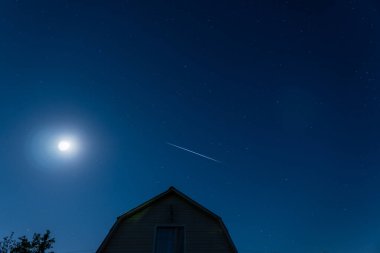 Bulutsuz, mavi, yaz gecesi. Starlink 'in tükürük hokkası evin üzerinde aya karşı uçuyor. Gecenin karanlığında, yıldızlı gökyüzü.