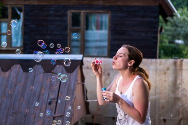 Yazın, eski ahşap evlerin arka planına sabun köpükleri üfleyen bir kız..